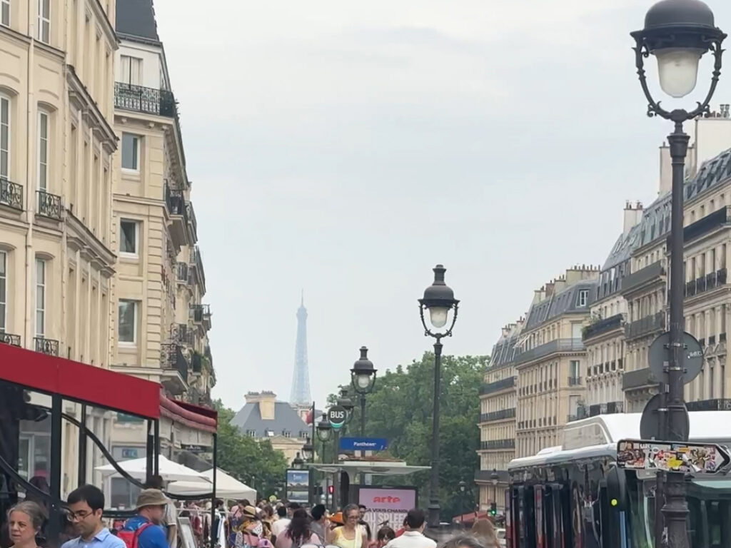 Crowded street with various people walking and standing on the sidewalk, showcasing urban life and activity.